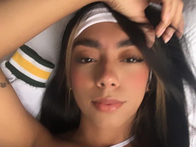 Alle69 with brown hair, white headband, golden earrings, pinkish blush, relaxed expression, hand in hair, subtle smile, slightly parted lips, soft lighting, tattoo on left arm.