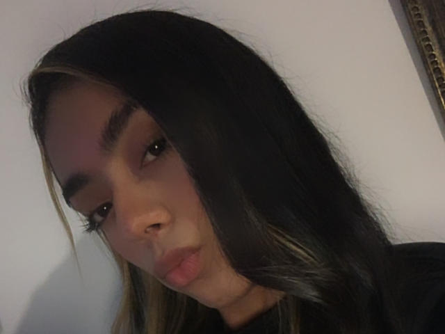 Alle69 has long black hair with blonde highlights, full eyebrows, and pouty lips. She's looking down with a slightly serious expression. Her nose is small and her skin is tan.