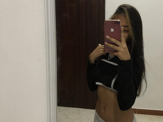 Alle69 with long brown hair, holding a pink iPhone, wearing a black top lifted to show her small tits and a grey sports bra, standing in front of a mirror.