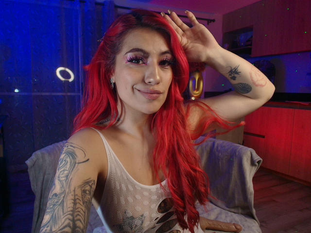 EvaMendex with bright red hair, wearing a white mesh top, showing tattoos on arms, smiling with hand in hair, blue and red lights, in a cozy room.