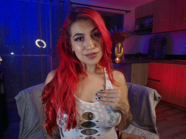 EvaMendex with bright red hair, wearing a white mesh top revealing her tits, has a hand on her chest, smiling, in a dimly lit room with blue and pink lights.