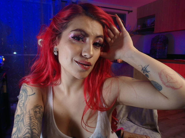 EvaMendex with red hair, wearing a white tank top, showing tattoos on arms, and a nose ring, smiling with pink eyelid makeup, and a hand in her hair.