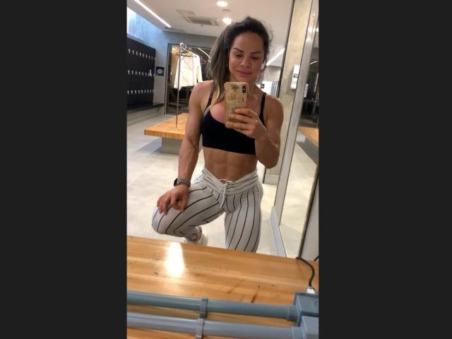 MiaBansolin, muscular, wearing a black sports bra and white striped pants, kneeling in a store, taking a mirror selfie, showing her toned abs and a small, firm butt.