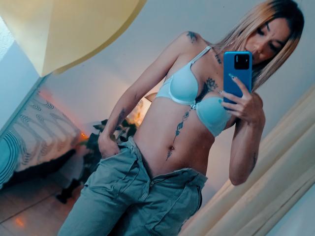 Veroo6g stands in a room, taking a mirror selfie. She's wearing a light blue bra that accentuates her small to medium-sized breasts and loose green pants. Her tattoos, including a large one on her lower abdomen, are visible. She has blonde hair and holds a blue phone in her right hand. The image is slightly blurred, with a yellow curtain in the upper left and a bed with a white blanket in the background. The room has a warm, soft light.