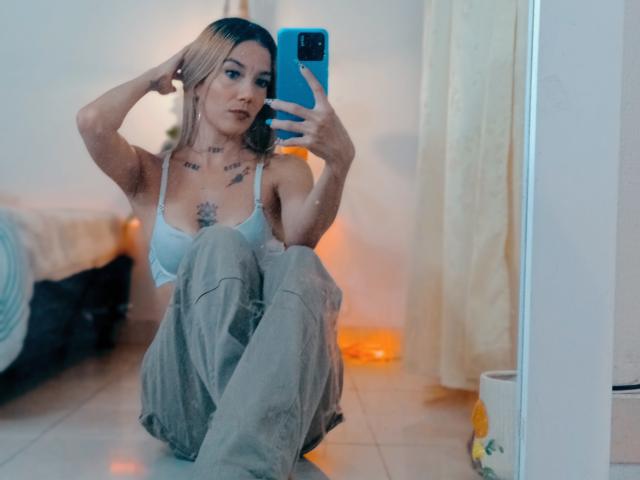 Veroo69 with blonde hair, white bra, and gray pants, sitting on floor, taking mirror selfie with blue phone, tattoos on chest, hands in hair, reflection shows bed and curtain.