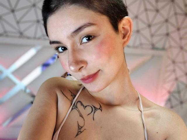 IsabellaHallad with short brown hair, slight pink blush, small tattoo of heart and lines on right shoulder, wearing a beige halter top, smiling softly at camera.