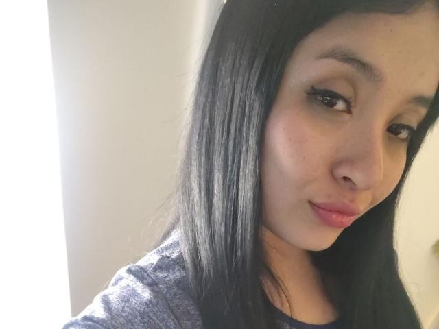 LunaBellaHotty has black straight hair, brown eyes, and a slight smile. She's wearing a gray shirt, and her pink lips are slightly parted. She's looking at the camera.