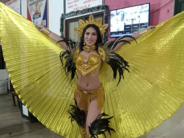 SofiaCumness in a gold and black feathered costume, arms spread wide, showing off shiny yellow wings and a glittery top with matching bottom, her tits and ass visible.