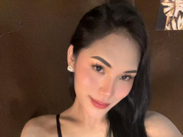SofiaCumness has black hair, thick eyebrows, red lipstick, and is smiling with a subtle, sexy look. She's wearing diamond earrings and a black strap top.