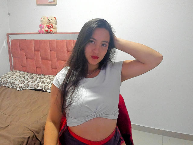 VenusSweetPalmer with long black hair, wearing a white cropped shirt, exposing her small tits, and red pants, hand in hair, sitting on bed with teddy bears in background.