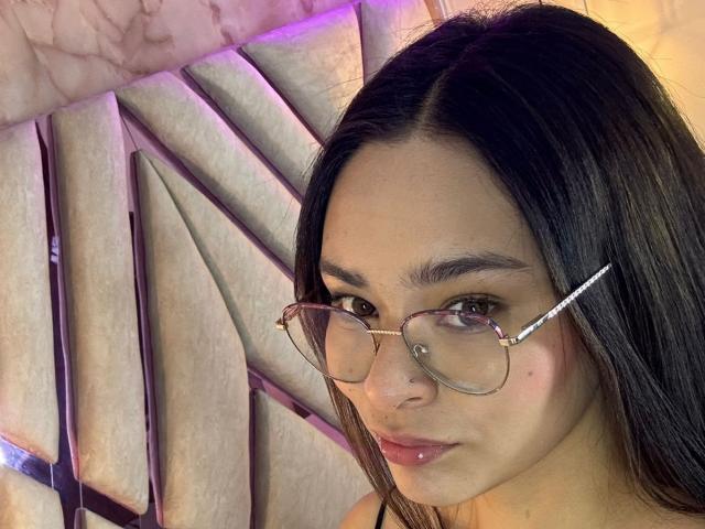 IsaDoux with medium brown skin, dark straight hair, wearing glasses, and pinkish lipstick, has a small mole on her right cheek, looking at the camera with a slight smile.
