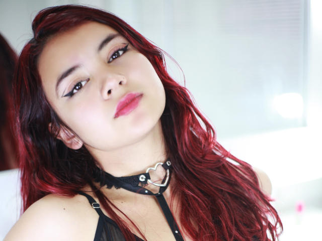Keittyx with red hair, black eyeliner, pink lips, wearing a black choker with a heart ring, looking up with a slight smile, showing her cute face and tits.