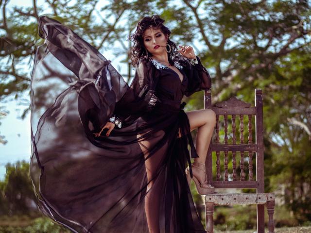 RenataRivas with black sheer dress, flowing sleeves, and deep cleavage, sitting on wooden chair, one leg up, wind blowing through hair, forest background, sultry gaze, high heels.