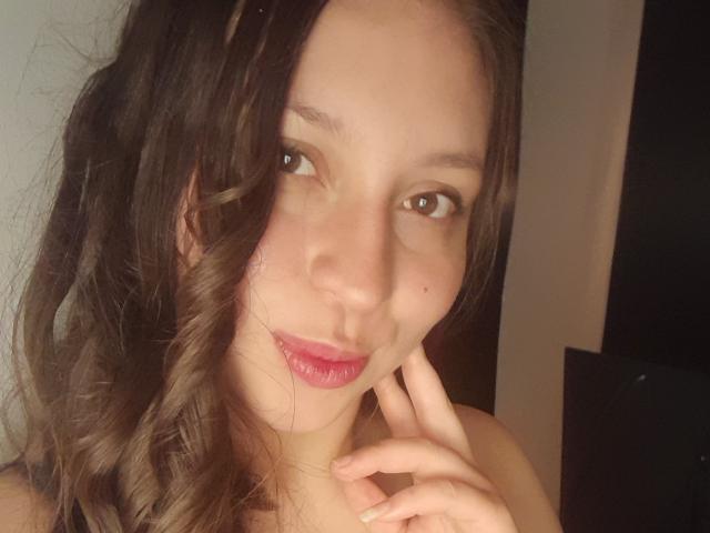 Malaika0203 has wavy brown hair, light skin, brown eyes, pink lipstick, and a small beauty mark on her left cheek. She's touching her chin with her right hand.