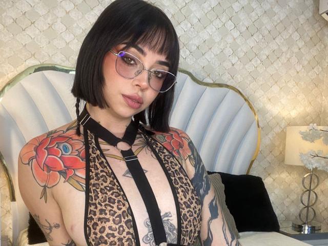 NaiaWhisper with a bob, glasses, leopard halter, large tits, tattooed arms, and red rose tattoos, looking at the camera. Bedroom with a white, gold-accented headboard.