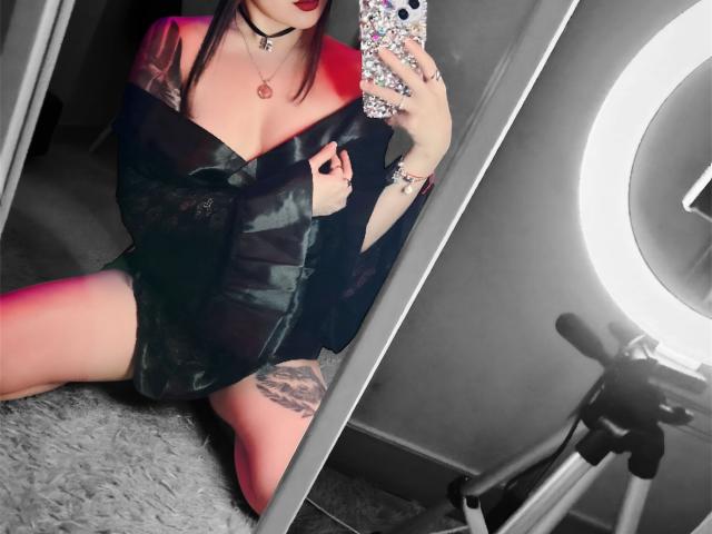 YukiiTyan with red lipstick, black off-shoulder shirt, and visible tattoo on thigh, kneeling, taking mirror selfie with phone, ring light on right, and bicycle in background.