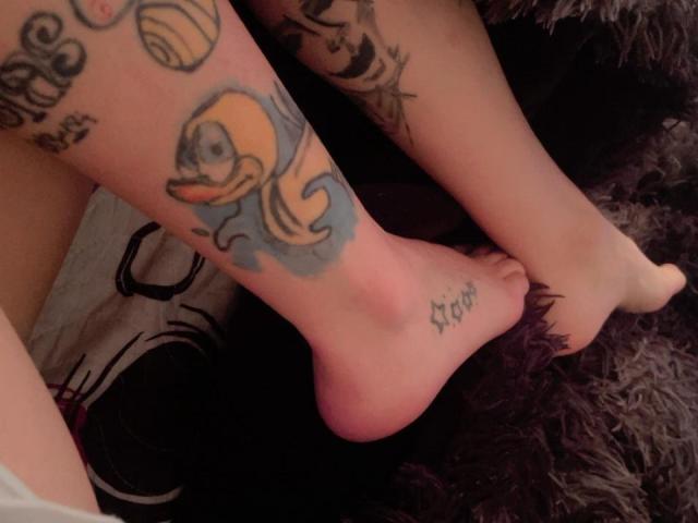 LaurenEvie69's tattooed foot with a blue and yellow cartoon duck, her other foot with a "Sick" tattoo, and black furry texture beneath. Her bare, pale skin is visible.