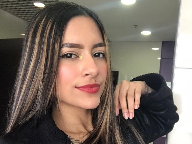 AfroditaWild ha la pelle marrone media, lunghi capelli castani lisci con riflessi biondi, sopracciglia nere, rossetto rosso e indossa un maglione nero e peloso, appoggiata a un muro bianco..