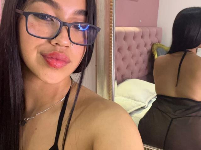 Nahomys with glasses, pink lipstick, and long black hair, wearing a sheer black top, showing her back and ass in a mirror reflection. She's smiling slightly.