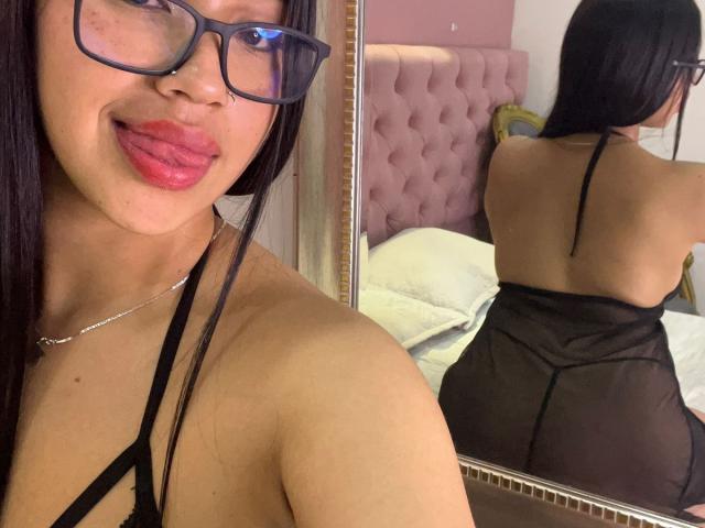 Nahomys with black hair, glasses, and red lipstick, wearing a sheer black halter top, showing her back and ass in a mirror selfie, in a bedroom.