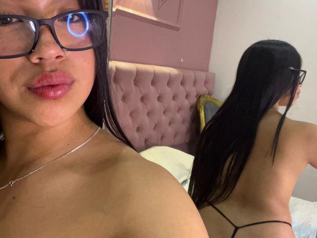 Nahomys with glasses, pink lips, and a necklace, facing camera, showing bare shoulders. Reflection in mirror shows her back, long black hair, and black thong.
