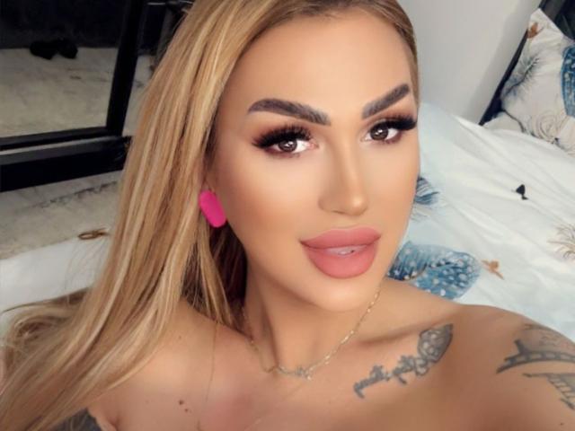 NicolSNaya has blonde hair, thick eyebrows, long eyelashes, pink lips, and pink earrings. She's topless with tattoos on her shoulder and chest, wearing a gold necklace.