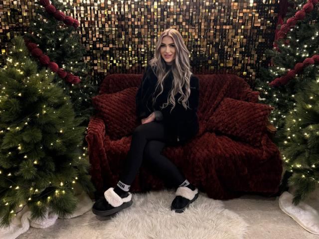 NicolSNaya with long blonde hair, black outfit, black tights, and black/white slippers, sitting on a red velvet couch between two decorated Christmas trees.