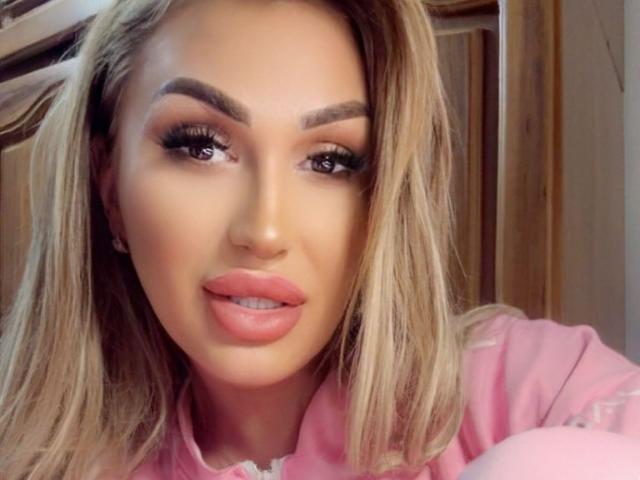 NicolSNaya with blonde hair, full lips, pink lipstick, and thick eyebrows, wearing a pink shirt, showing off her sexy face and big ass.