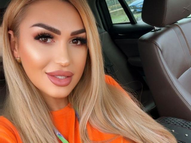 NicolSNaya has blonde hair, brown eyes, and full lips with pink lipstick. She's wearing an orange top, looking into the camera with a slight smile.