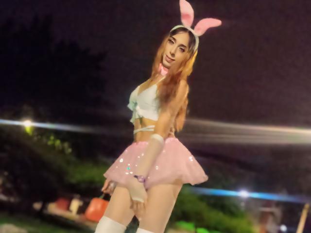 DanielaWalker with pink bunny ears, white crop top, pink sequined skirt, white thigh-highs, and long brown hair, standing at night, showing her ass and tits.