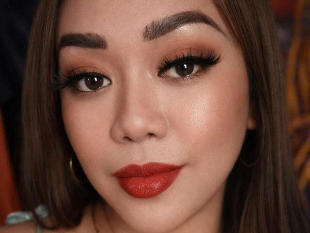 Close-up of BigSauvageQueenTs. She has smooth, light brown skin, full eyebrows, and brown eyes with long, thick black eyelashes. Her red lipstick stands out against her slightly shiny, even complexion. She has long, straight brown hair, and is wearing small, gold hoop earrings. The image focuses on her face, with her slightly smiling lips. The lighting is even, highlighting her features. The overall style is realistic and polished.