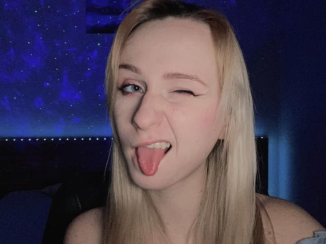 FlyFreedom has blonde hair, fair skin, and is winking with her tongue out. She has visible eyeliner and a cute, playful expression. Her nipples are slightly visible.