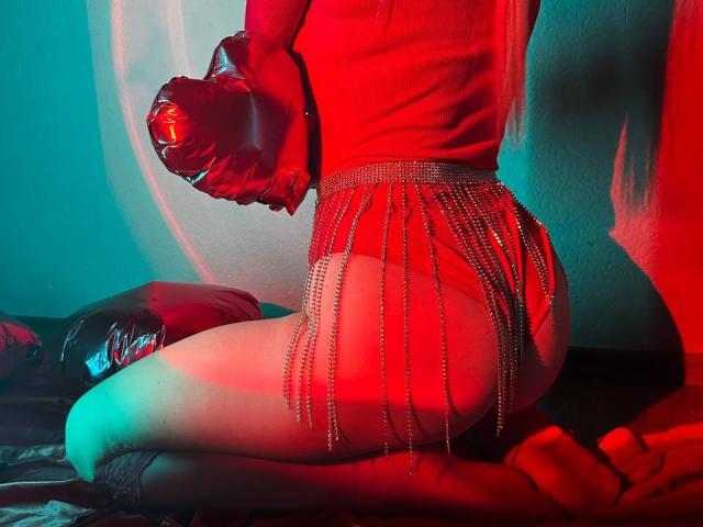 BalletSwan's red top, shiny butt, and sparkly chain skirt, kneeling on a bed with a black pillow, wearing black socks, under red and blue lighting.