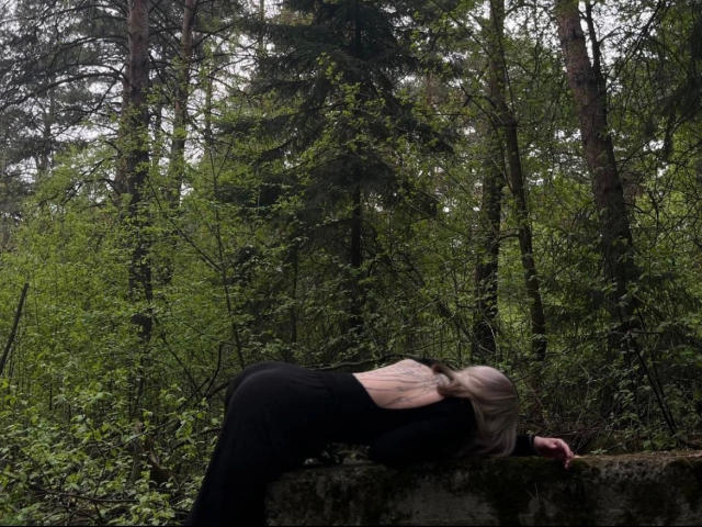 Unknown-hot with long blonde hair, black top, and black pants bends over a mossy stone, her ass raised, hands on the ground, in a green forest.