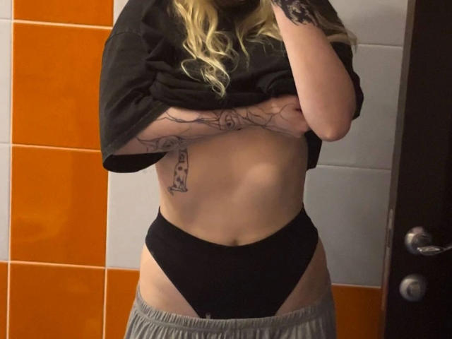 Unknown-hot with blonde hair lifting a black shirt, exposing her small tits and black panties, showing a tattoo of a flaccid penis on her side.