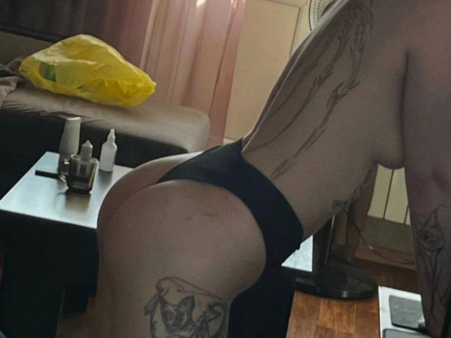 Unknown-hot with tattoos, leaning forward, wearing black thong, showing ass, small tits, and tattoos on side and thigh, in dimly lit room, with yellow bag on brown couch.