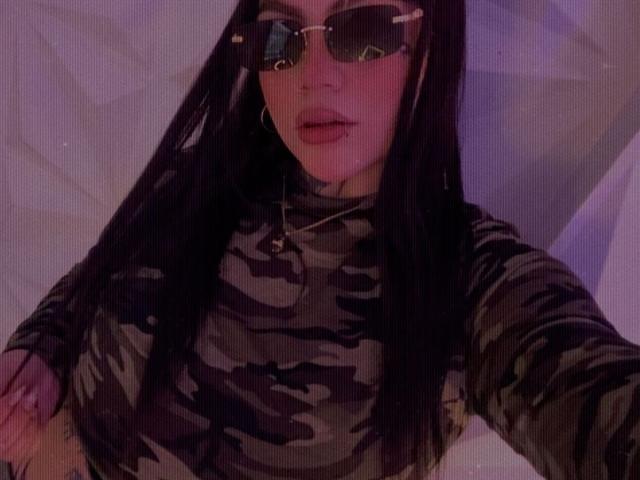 TessaMagic with long black hair wears reflective sunglasses and a camo-patterned, long-sleeve shirt. Her lips are slightly parted with a subtle pink lipstick. She has a small tattoo on her left forearm. She wears a small necklace with a cross pendant. The image has a purple tint. Her right hand is slightly raised. The composition is a close-up selfie.