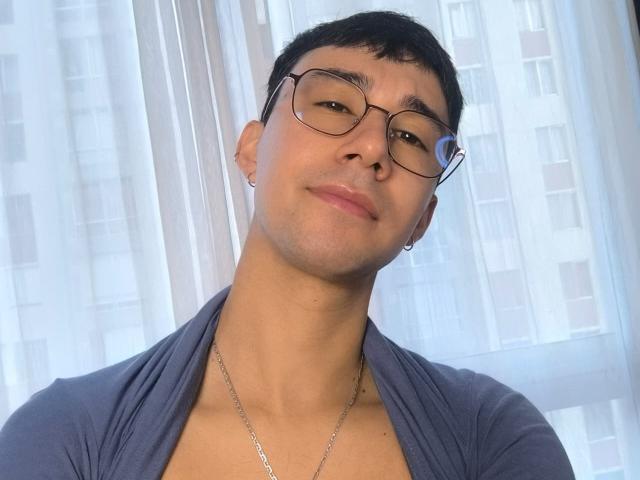 OliverHot69 with glasses, short black hair, slight smile, wearing a blue shirt with a V-neck, silver necklace, and small earrings, looking slightly up.