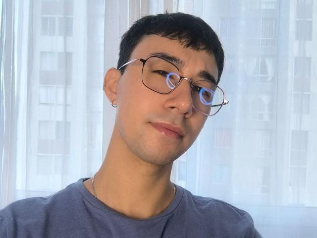 OliverHot69 with short black hair, glasses, and a slight smile, wearing a blue shirt, has a small earring in his left ear, and is looking directly at the camera.