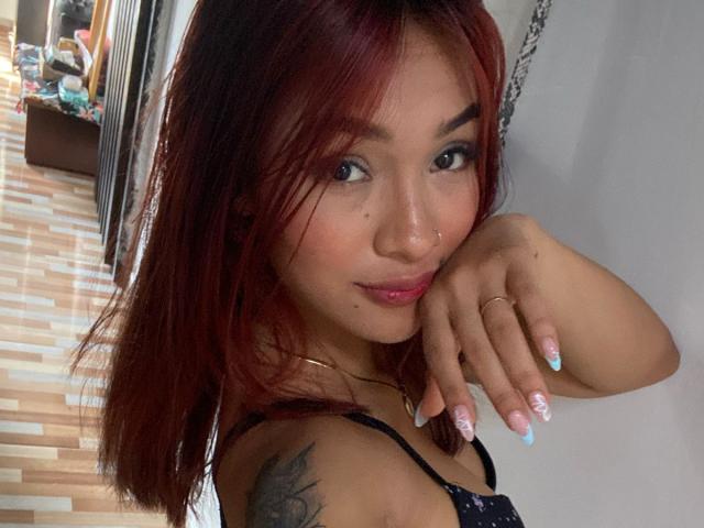 SashaHans, avec ses cheveux roux, ses yeux verts et un piercing au nez, souriante, montrant ses fesses avec une culotte noire à fines bretelles et un tatouage sur l'épaule, avec des ongles peints en bleu..