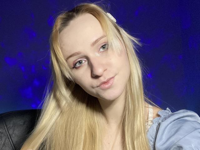 FlyFreedom has blonde hair, light skin, and blue eyes, wearing a light blue shirt with a white hair clip, and a slight, relaxed smile. Her tits are small.