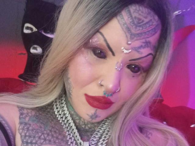 VenusHottt with blonde hair, tattoos on forehead and neck, red lipstick, nose piercings, chain necklace, and glitter on face, looking at camera with a slight smile.