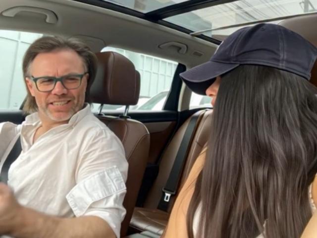 ViktorAndPenny with long brown hair, wearing a black cap, sits in car's brown leather seat. ViktorAndPenny with glasses, white shirt, smiles, driving.