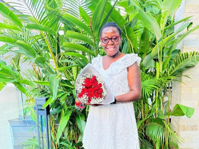 WillaSara è in piedi, con un bouquet di fiori rossi e bianchi, indossa un abito bianco in pizzo, occhiali e guanti grigi, e dietro di lei ci sono piante verdi. Sorride dolcemente..