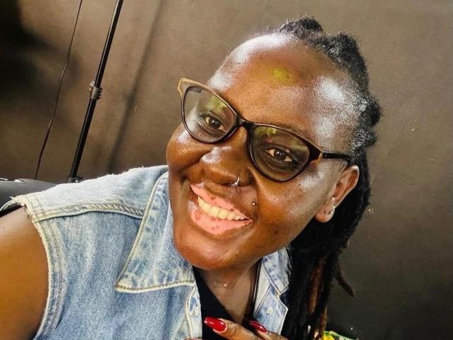 WillaSara, à la peau foncée, souriante, portant des lunettes noires, un gilet en jean, un anneau au nez et des ongles rouges, a les cheveux tressés et regarde l'appareil photo. Son front brillant présente une marque jaune..