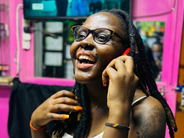 WillaSara, donkerhuidig met dreadlocks, draagt een bril, glimlacht met haar tong uitgestoken, houdt een telefoon op haar oor, heeft gekleurde nagels, tatoeages, en op de achtergrond is een roze muur te zien, en draagt een zwart tanktop..