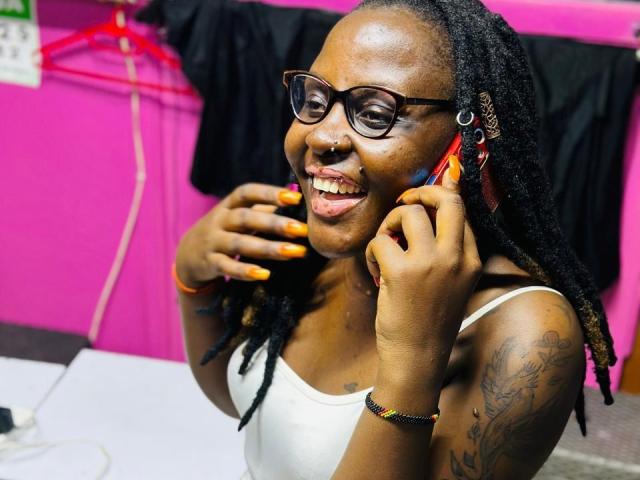 WillaSara, met donkere huid en gevlochten haar, draagt een bril en een wit tanktop, ze glimlacht met oranje nagels, en heeft een neusring en tatoeages, ze houdt aan haar oor..