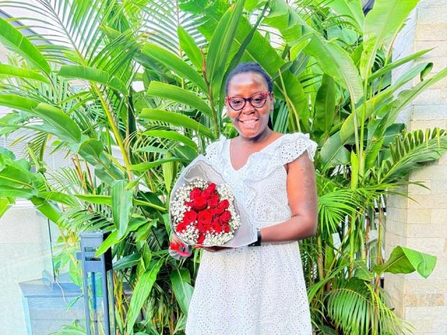 WillaSara with medium brown skin, glasses, and white dress, holding a bouquet of red and white flowers, standing in front of lush green banana leaves.