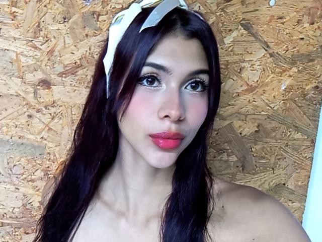 DanikaSerov has long, dark brown hair with a white bow on top. She has large, dark brown eyes with thick, black eyeliner and long lashes. Her skin is light with a slight pink blush on her cheeks. She has full pink lips. She is topless, with bare shoulders visible. The background is a textured, wooden surface. The image is a photograph with natural light highlighting her face. Her expression is neutral.