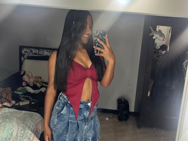 NaomiRios with long black hair, smiling, wearing a red tied-up top showing her tits and blue denim high-waisted jeans, taking a mirror selfie in a bedroom.